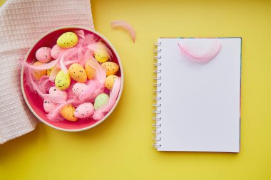 Yaratıcı Paskalya not defteri sarı arka planda. Madalyalı Paskalya: Renkli yumurtalar ve pembe tüylü pembe kase.