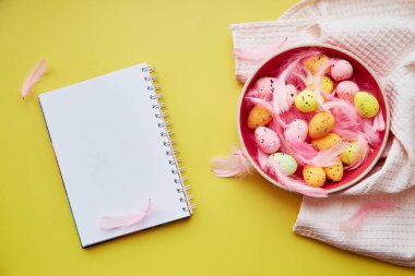 Creative bright Easter notepad mock up on yellow background. Decorated Easter: pink bowl with colorful eggs and pink feathers