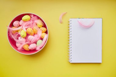 Minimalist Easter notepad mock up on yellow background. Pink bowl with colorful eggs and pink feathers. Copy space.