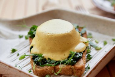 Vegan eggs benedict dish with wholegrain bread, creamy plant-based hollandaise sauce, smoked tofu, young onion, parsley, greens, pea sprouts