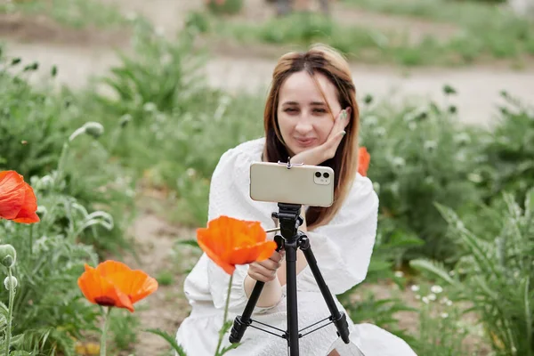 Beyaz elbiseli gülümseyen milenyum kızı gelincik tarlasında tripod ve telefon kullanarak video kaydediyor. Blogculuk yaşam tarzı. Modern teknolojiyi kullanarak