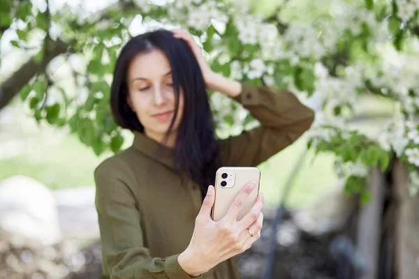 Woman is making selfie by camera phone under blooming tree. Selective focus
