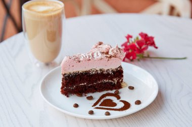 Meyveli çikolatalı pasta ve açık masa üzerinde duble latte olan bir bardak. Kafe terasında pembe çiçek süslemeleri. Atmosferik kahvaltı