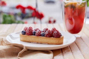Terasta ahududu ve yaban mersinli Fransız şekersiz tart. Ahududu ferahlatıcı- portakallı limonata, dışarıda nane ve kamış kamışı ile ahşap masada.