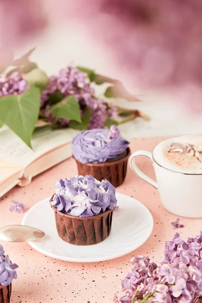 Aesthetic sprimg background. French purple floral cupcakes using trend Dreamy Escapism with lilac flowers and book. Leisure and relax coffee time.
