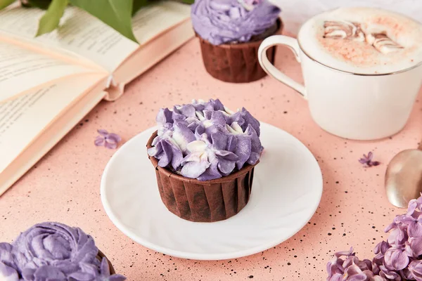 Aesthetic lifestyle.Book and purple trendy floral cupcakes with cup of coffee. Violet sweet no sugar dessert among lilac flowers.