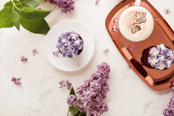 Aesthetic flat lay table setting. Purple french cupcakes on the golden tray with coffee cup and lilac flowers.