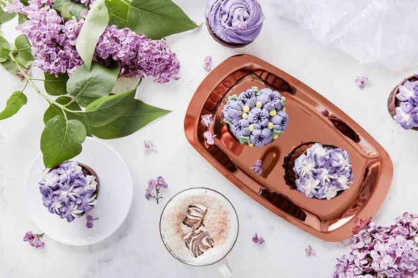 Feminine floral background. Purple french cupcakes. Desserts on the golden tray, coffee cup and lilac flowers flat lay