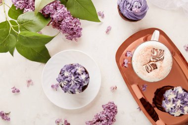 Çiçekli mor çörekler, erimiş marşmelovlu kahve fincanı leylak çiçeklerinin arasında düz bir zemin.