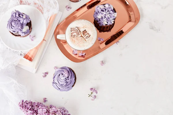 Purple aesthetics cupcakes flat lay, desserts on golden tray, coffee cup with melted marshmallow among lilac flowers background.