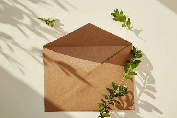 Craft paper open envelope mock up under sunny shadows with tiny leaves. Copy space.