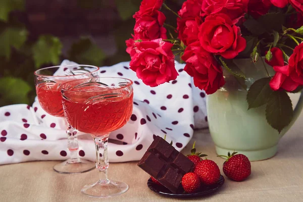 Glasses of red sparkling strawberry wine with roses outdoor, strawberry and chocolate. Aesthetic summer table settings. Romantic, precious moment concept
