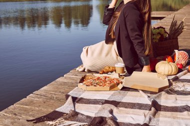  Sonbahar süslemelerinin arasında bir kadın, balkabakları iskelede piknik yapıyor. Sonbahar estetik piknik yemekleri: pizza, kahve ve kruvasan, sert gölgeler..