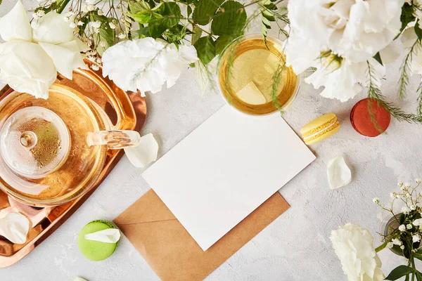 Aesthetic craft envelope with stationery card mock up for greetings, invitation, text among white flowers, macaroons and tea. Biophilic interior, cozy home. Copy space