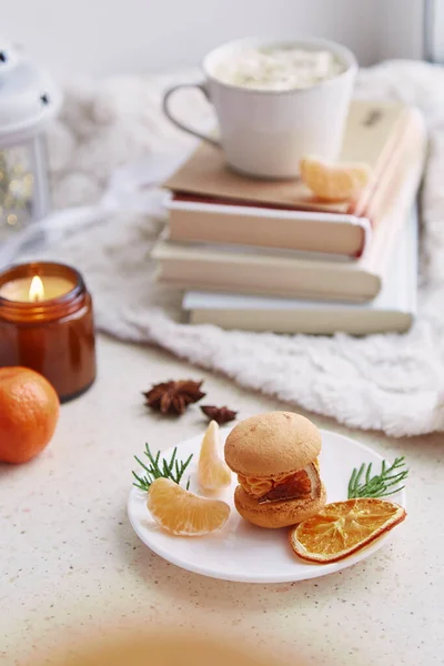 Aesthetics dessert bright orange macaroon, tangerines among books, candle and coffee. Festive table setting.