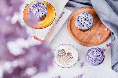  Estetik mor masa dekorasyonu. Kekler ve kahve fincanı kitaplarda, leylak çiçeklerinin arasında. Uzayı kopyala Suçlu zevk, değerli an.