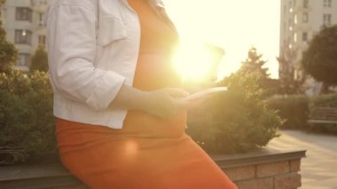 Tanımlanamayan hamile kadın kahve içiyor ve gün batımında akıllı telefon kullanıyor. Bekleyen kadın bankta oturuyor, cep telefonu kullanıyor ve rahatlıyor.