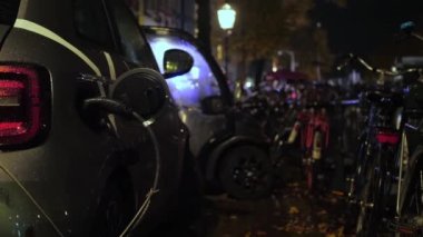 Elektrikli araba, arka plandaki EV şarj cihazına bisikletlerle bağlı. Gece vakti, yağmurlu bir havada, halka açık bir istasyonda araba şarj oluyor.