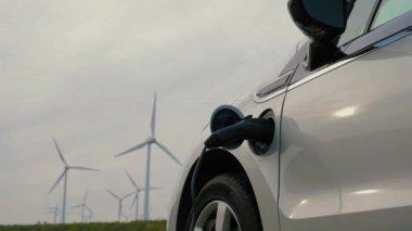 Elektrikli araç şarj olurken, rüzgar türbinleri arka planda yenilenebilir enerjiyi temsil ederek sürdürülebilir enerji ve yeşil teknolojiyi sembolize ediyor. Güç bağlantısı EV kopyalama alanı