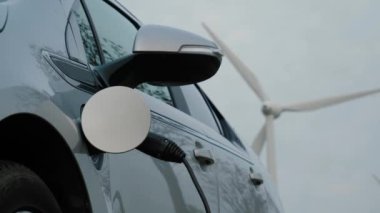 Yenilenebilir enerji, sürdürülebilir enerji ve yeşil teknolojiyi sembolize ediyor. Elektrikli araç şarj olurken rüzgar türbinleri arka planda dik duruyor. Güç bağlantısı EV kopyalama alanı