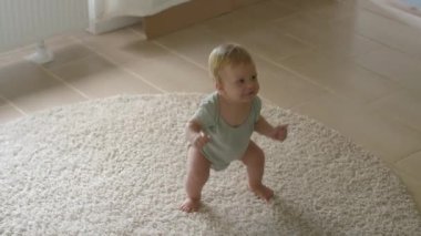Neşeli bebek yumuşak, rahat bir halının üzerinde duruyor, gururla yeni başarılarını gösteriyor. Sevimli küçük çocuk dans eder ve çömelir. Bebek ayakta durmayı yeni öğreniyor.