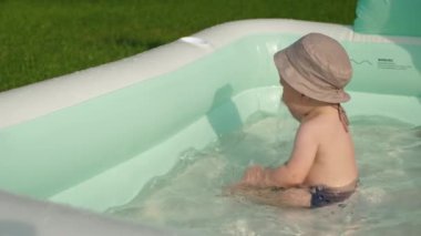 Şık güneş panama şapkalı mutlu çocuk şişme havuzda kameraya bakıyor, yaz eğlencesini somutlaştırıyor ve unutulmaz anılar yaratıyor. Bebek eğleniyor, arka bahçede su sıçratıyor, ağır çekim.