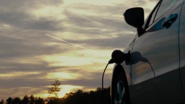 Doğada park edilmiş elektrikli araba, EV tip 1 kablosu bağlı ve şarj bataryası, arka planda gün batımı, zaman atlaması. Elektrikli araç kavramında yenilenebilir enerji