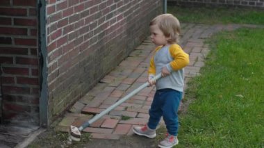 Neşeli çocuk kırmızı tuğla duvara karşı süpürgeyle oynuyor. Küçük çocuk yetişkinler için somutlaştırıcı fırça taşıyor. Bahçeyi keşfeden çocuk