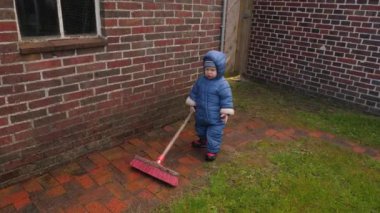 Parlak mavi yağmurluk giymiş neşeli küçük bir çocuk. Bebek mutlu bir şekilde tuğla yolu süpürüyor sağlam tuğla duvarın yanında, süpürgeli bebek yağmurlu bir günde çalışmaktan zevk alıyor.