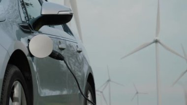 Elektrikli araç şarj olurken rüzgar jeneratörleri arka planda, sürdürülebilir enerji ve yeşil teknolojiyi sembolize ederek yenilenebilir enerjiyi temsil ediyorlar. Güç konnektörü EV, yakınlaştır