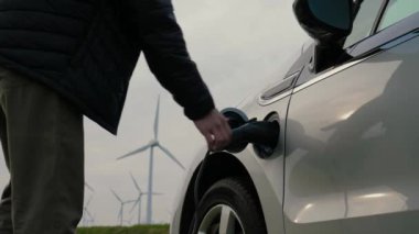 Tanımlanamayan kişi elektrik kablosunu rüzgar jeneratörlerinin yanında şarj olan elektrik arabasından ayırarak sürdürülebilir enerji ve yeşil teknolojiyi sembolize ediyor. Erkek eller güç bağlantısını EV 'den ayırır