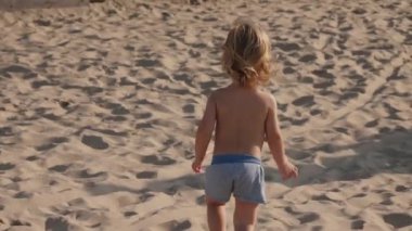 Neşeli çocuk kumlu sahilde coşkuyla yürüyor, güneşli günün güzelliklerine dalmış, tasasız özünü, çocukluk ruhunu ve yaz eğlencesini sergiliyor.