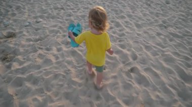 Parlak sarı gömlekli ve yeşil şortlu küçük bir çocuk kumsalda mutlu bir şekilde yürüyor, elinde annesi için mavi terlikler tutuyor, kumsalda güzel bir günün keyfini çıkarıyor.