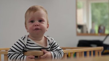 Yüzünde tuhaf bir ifade olan sevimli bebek tahta oyun parkında duruyor. Küçük çocuk sıcak ve rahat oyun odasında, herhangi bir çocuk için hazırlıyor.