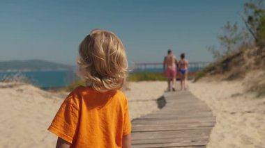 Güneşli bir günde, turuncu tişörtlü küçük bir çocuk plaja giden yolda heyecanlı bir şekilde yürürken, parlak renkli mayo giymiş iki yetişkin önlerinde yürüyüp deniz kenarındaki atmosferin tadını çıkarıyorlar.