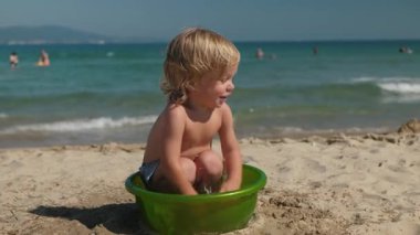 Kıvırcık saçlı küçük neşeli çocuk, açık mavi suların kıyıya kavuştuğu güneşli sahildeki yeşil havuzda oynuyor. Bebecik plajda güneşin tadını çıkarıyor.