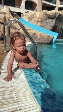 Havuz kenarında neşeli bir çocuğun oynadığı, ferahlatıcı suyun ve sıcak güneşin tadını çıkardığı, keyifli bir tatil sırasında yavaş çekimde dikey bir video. Rahatlatıcı bir ortamda çocukluk sevinci