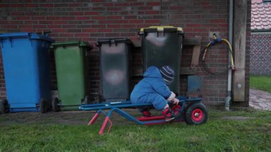 Kışlık palto giymiş küçük bir çocuk çimenli arka bahçeye yerleştirilmiş küçük bir yük vagonunda rahat bir şekilde oturur. Tuğla evin yanında huzurlu bir anın tadını çıkarır.