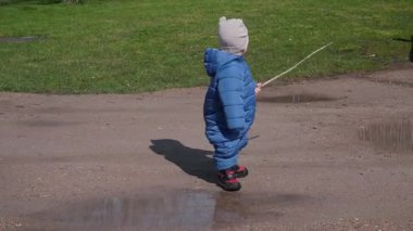 Neşeli küçük bir çocuk, rahat mavi bir kar elbisesi giymiş, sopayla su birikintilerini keşfediyor, kameraya bakıyor. Bebek, açık hava eğlencesinde neşeli çocukluk, merak ve eğlencenin özünü somutlaştırıyor.