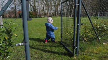 Güneşli bir bahar gününde, parlak mavi takım elbiseli küçük bir çocuk ilginç bir şekilde etrafı yemyeşil çimenler ve renkli bitkilerle çevrili büyüleyici bir bahçe kapısını keşfeder.