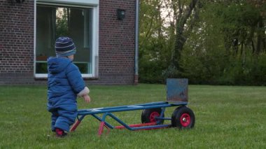 Kışlık palto giymiş küçük bir çocuk, bir el arabasını coşkuyla yemyeşil bir arka bahçeye itiyor, dışarıda geçirdiği huzurlu anın tadını çıkarıyor, büyüleyici, rahat tuğlalı bir evin yanında.