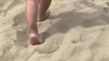 Tanınmayan çocuk kumlu bir sahilde neşeyle çıplak ayakla yürüyor, altın kumda ayak izleri bırakıyor, mutluluk ve macera dolu sahilde geçirilen bir günün saf mutluluğunu yakalıyor, ağır çekimde.