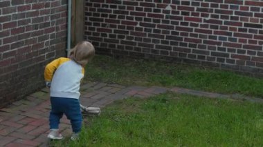 Bahçeyi keşfeden çocuk. Neşeli çocuk kırmızı tuğla duvara karşı süpürgeyle oynuyor. Küçük çocuk yetişkinlere yardım etmek için fırça taşıyor.