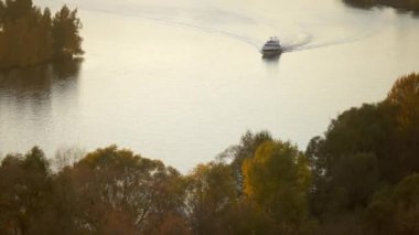 Sakin bir nehir boyunca gün batımında seyreden beyaz bir motor yatı, yoğun bir şekilde sulara yansıyan altın sonbahar yapraklı ağaçlarla kaplanmış. Altın sonbahar manzarası konsepti