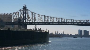 New York, Roosevelt Adası Köprüsü. Köprü altından New York 'taki binaların arka planına karşı köprüden geçen arabalarla nehir boyunca büyük bir mavna yüzüyor. New York, Amerika - 02 Mart 2020.