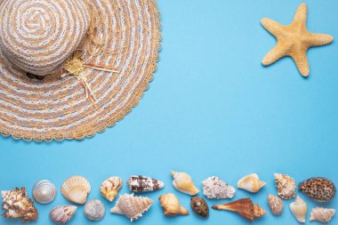 Yaz ve tatil düzlüklerinde bir kadın hasır şapka, bir yıldız balığı ve mavi arka plandaki resmin alt tarafında çeşitli deniz kabukları yatıyordu..