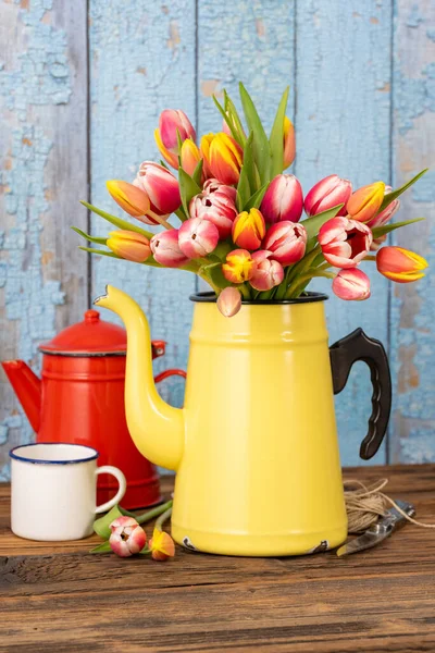カラフルなチューリップの花束を持つエナメルコーヒーポットは素朴な木製のテーブルの上に立っています その後ろに別のエナメルコーヒーポットとコーヒーマグカップ — ストック写真