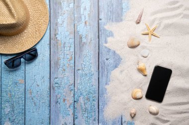 Yaz, sahil ve bedava mesaj alanı olan tatil konsepti. Üst Manzara. Hasır şapkalı, güneş gözlüklü, akıllı telefonlu, çeşitli deniz kabuklu ve eski mavi ahşap arka planda kumlu düz bir düzen.