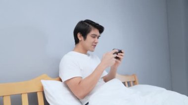 Tatil konsepti 4k Çözünürlük. Asyalı adam yatak odasında mobil oyun oynuyor. online oyun eğlencesi.