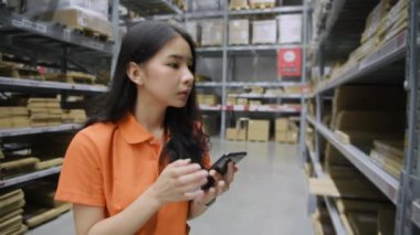 4k Kararlılığın iş konsepti. Asyalı bir kadın bir depoda cep telefonundan yapılan başvuru ile ürünleri kontrol ediyor..